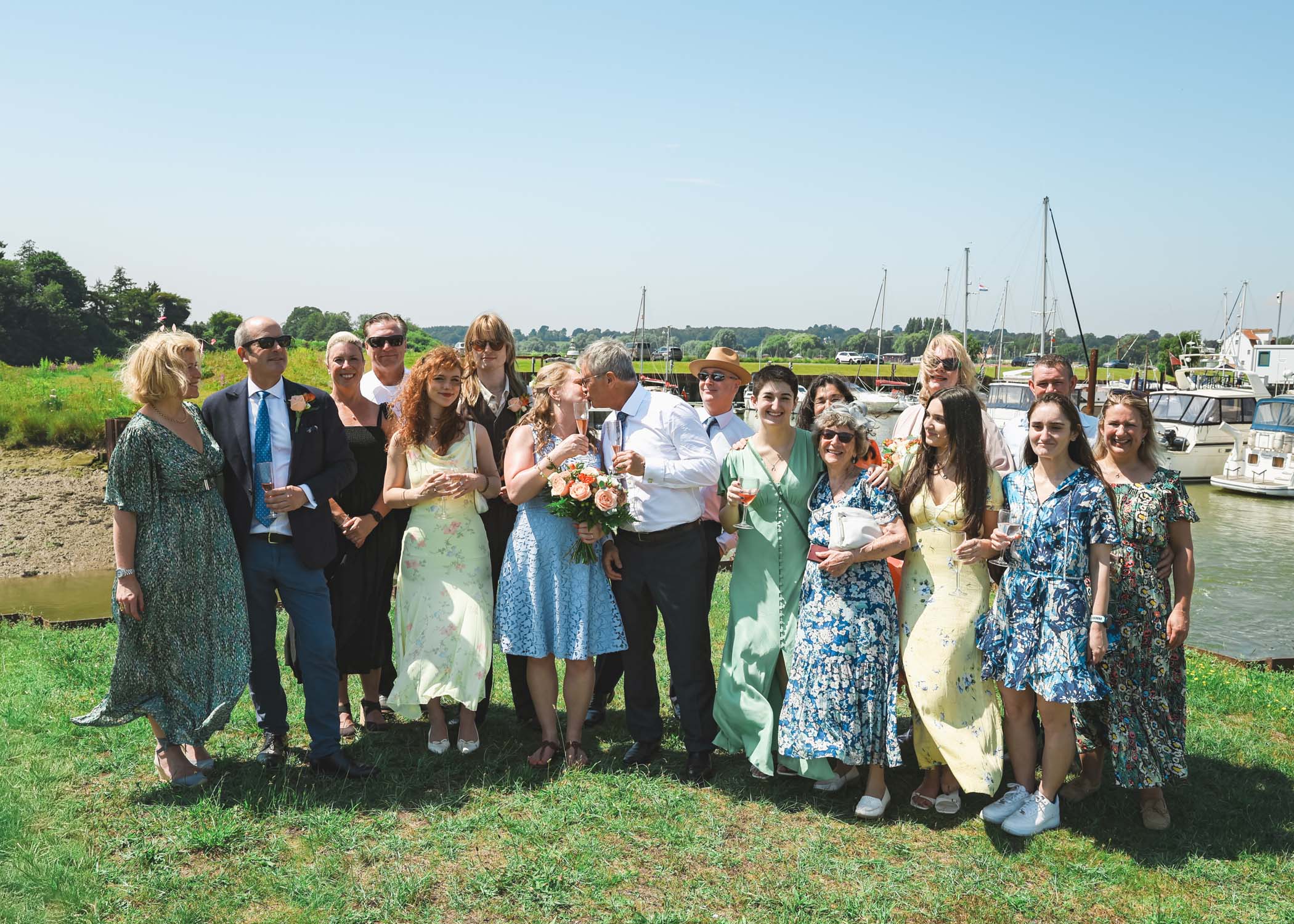 A group photo with the bride and groom kissing in the middle on the penisula at woodbridge marina on a wedding day in Woodbridge captured by Suffolk Wedding Photographer Hayley Denston Photography
